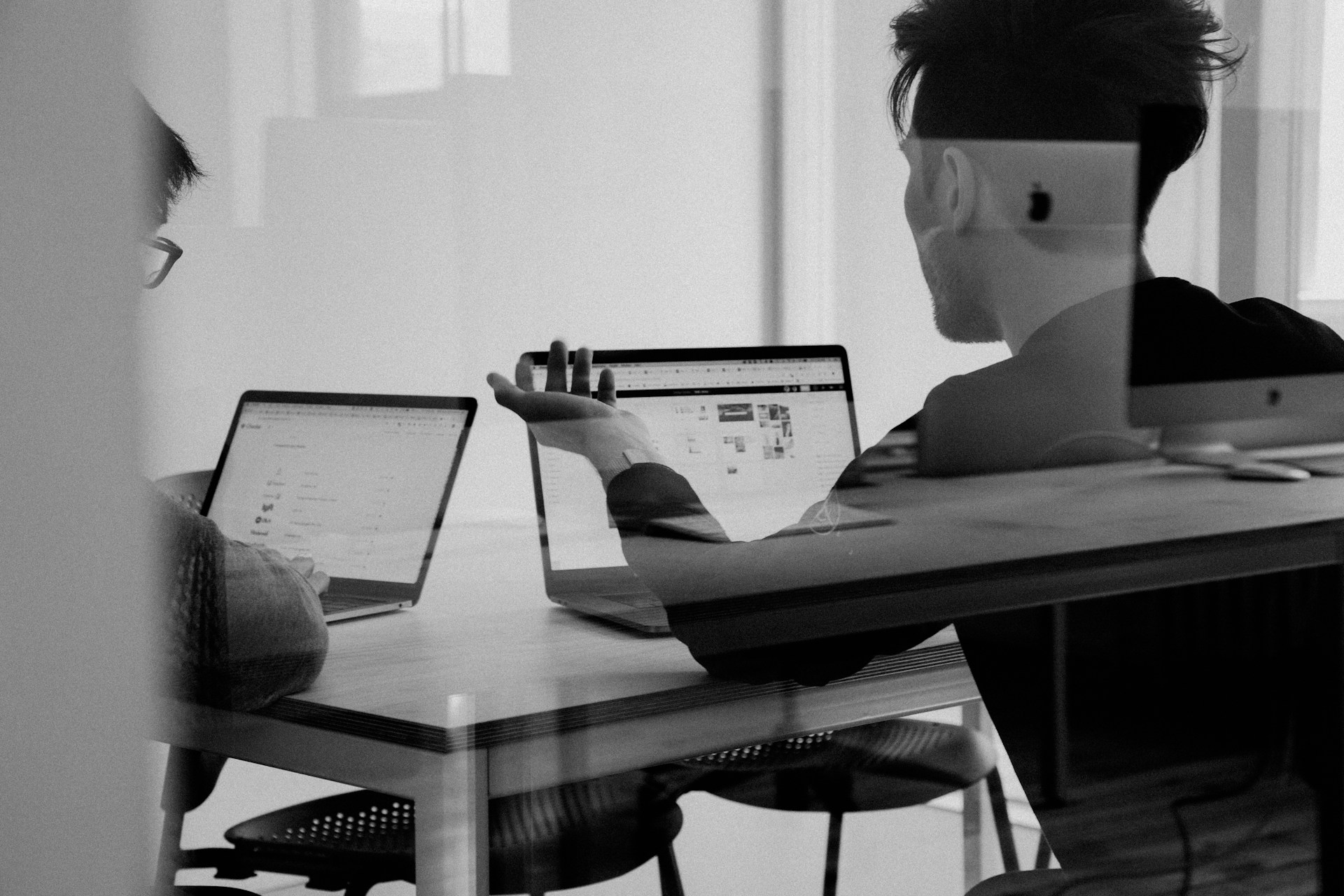 A photo of two people at a table looking at separate laptops. The photo is taken through a glass pane.
